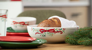A festive table setting with green and red plates, a bowl with holiday decorations holding bread covered by a white napkin, and a sprig of pine in the foreground. The bowls have a red ribbon and pine design.
