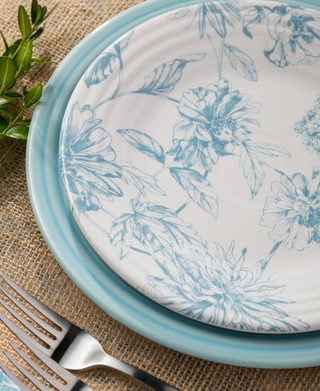 A close-up of the Fiesta Botanical Floral Sky Classic Rim 9 Inch Luncheon Plate, made in the USA, with a matching dinner plate set on burlap with cutlery and a sprig of greenery beside them.
