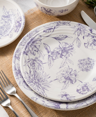 Two stacked Fiesta Botanical Floral Lavender Classic Rim 9 Inch Luncheon Plates rest on a burlap placemat with a fork and knife to the left. Matching dishes with the same delicate purple floral pattern are partially visible nearby.