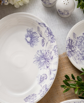 The Fiesta Botanical Floral Lavender 11 5/8 Inch Medium Oval Serving Platter rests on a table with a matching plate, green leaves, and a textured placemat, while salt and pepper shakers appear in the background.