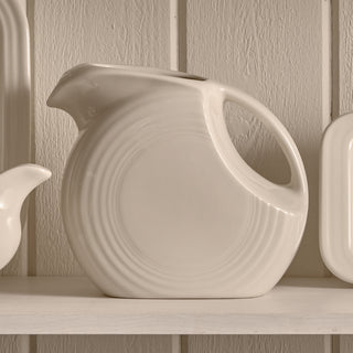 The Fiesta 67 OZ Large Disk Pitcher by Fiesta, crafted as lead-safe china, features a cream color, rounded Art Deco design, and curved handle. It sits on a shelf against a light wooden background.
