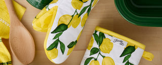 A lemon-patterned oven mitt and pot holder lie on a wooden surface next to a wooden spoon, a yellow towel, and a green baking dish.