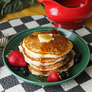 A stack of pancakes with butter and syrup sits in a green Fiesta Coupe 8 1/2 Inch Luncheon Bowl (26 oz), surrounded by strawberries and blueberries, with a fork and red syrup pitcher on a checkered placemat nearby.