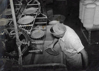 A worker wearing a cap dips a plate into a vat, with other plates on racks nearby in a ceramics factory. Stacks of finished plates are visible in the background.