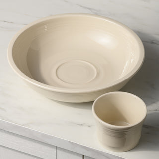 The Fiesta 2-Piece Chip and Dip Set (12 7/8 inch), featuring a cream color and glossy finish, includes a large bowl with a subtle circular pattern and a matching cup—perfect for entertaining—displayed on a white marble countertop.