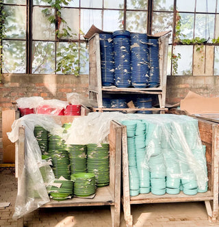 Stacks of colorful bowls—green, light blue, red, and navy blue—are sorted on wooden carts, partially covered with plastic sheets, inside a sunlit room with brick walls and large windows with climbing plants.