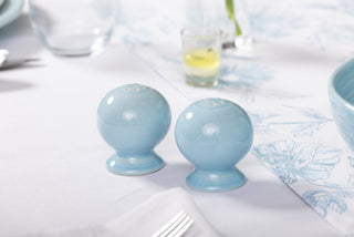 Two round, light blue Fiesta 2 5/8 Inch Salt and Pepper Shaker Set shakers sit side by side on a white tablecloth, with other tableware and a small yellow drink in the blurred Art Deco background.