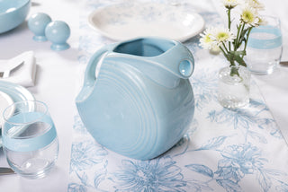 A Fiesta 67 OZ Large Disk Pitcher in light blue sits on a white and blue floral tablecloth, surrounded by matching Fiesta lead-safe china dishware, a glass of water, salt and pepper shakers, and a vase with white and yellow flowers.