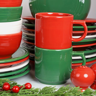 A stack of red, green, and white dishes and bowls, with red and green mugs in the center, next to a red ornament and greenery with red berries, evoking a festive, holiday theme.