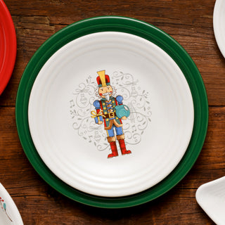A white plate with a green rim features an illustration of a toy soldier in a red, blue, and gold uniform holding a drum, set against a festive, decorative background on a wooden table.