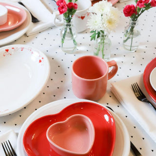 A table set with Fiesta Retired 7 OZ Heart Shaped Ramekins (4 1/2") in red and pink, a pink mug, white plates, small vases of red and white flowers on a white tablecloth with black hearts. Utensils rest on white napkins.