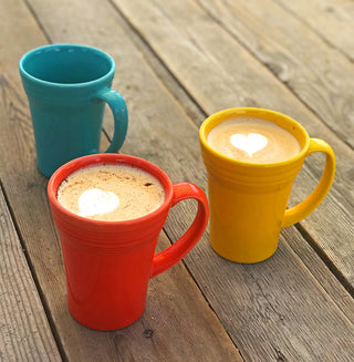 Three Retired Fiesta 18 OZ Bistro Latte Mugs by Fiesta—blue, red, and yellow—rest on wood. The red and yellow mugs contain coffee with heart-shaped foam art; all are dishwasher safe for easy cleaning.