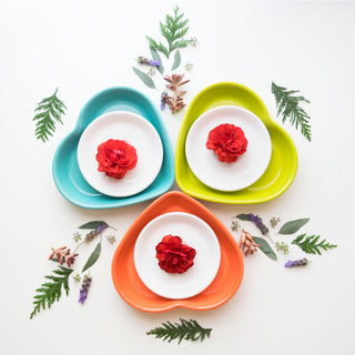 Three Retired Fiesta 7 1/2 Inch Medium Heart Shaped Bowls (17 OZ) in teal, yellow, and orange by Fiesta are arranged in a triangle on a white surface, each holding a white plate with a red flower, surrounded by green leaves and small flowers.