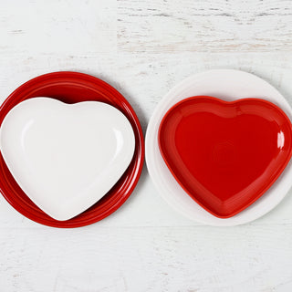 Two retired Fiesta 9-inch heart-shaped plates are displayed: a white heart plate on a red round plate to the left, and a red heart plate on a white round plate to the right, all by Fiesta on a white wooden surface.