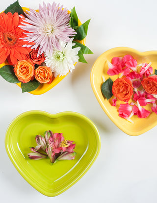 Three Fiesta Retired 7.5" Medium Heart Shaped Bowls (17 oz)—two yellow, one green—made in the USA from lead-safe china, are filled with orange roses, pink and white chrysanthemums, and pink alstroemeria on a white background.